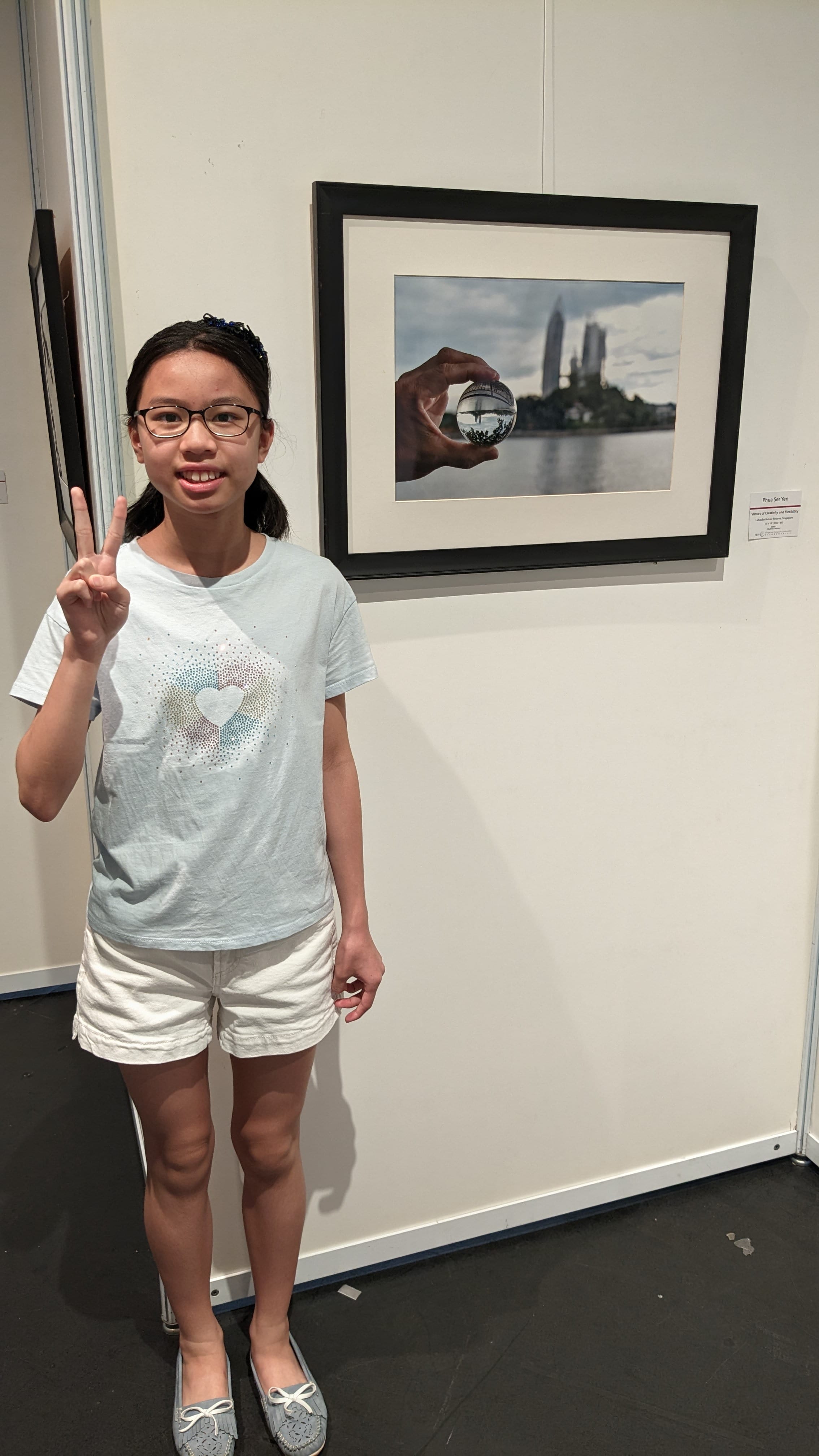A young girl with glasses makes a peace sign beside a framed photo of a hand holding a glass ball with a cityscape.
